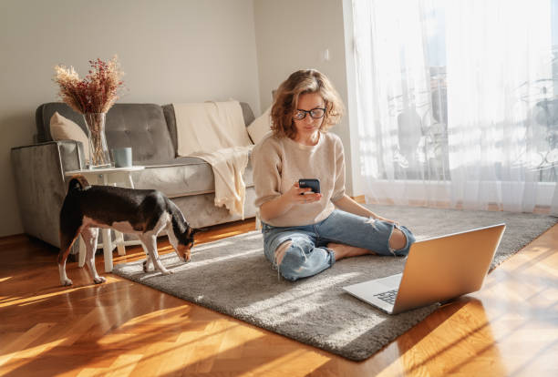 Pretty curly happy young woman sitting at home on the floor in front of laptop with her pet dog working and learning online with smartphone in hands stock photo
