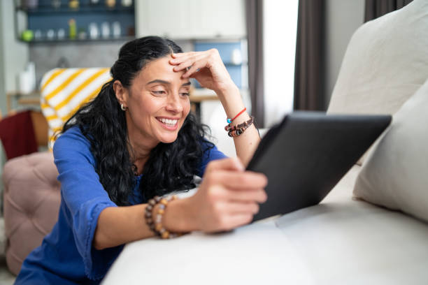 Pretty curly woman sitting on the sofa, holding a digital tablet, smiling and having online communication stock photo