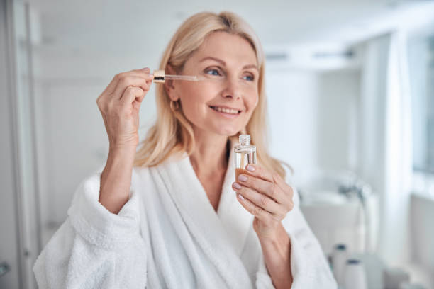 Pretty elegant woman using natural cosmetic product for hydrated and facial healthy derma Close up portrait of charming woman holding bottle with dropper near face while applying cosmetic facial oil on face with pipette in bathroom face oils stock pictures, royalty-free photos & images