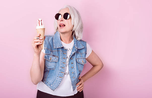 Pretty fanny senior woman holding icecream. My passion. Emotional beautiful senior woman holding an icecrem and licking lips while standing against isolated pink background. baby boomers stock pictures, royalty-free photos & images