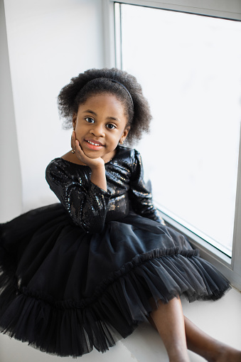 Portrait of pretty female kid in black stylish dress sitting near window in white studio and looking at camera. Cute face of african american petite ballerina.