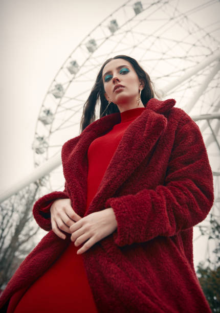 Pretty girl against Ferris wheel. Retro (vintage) portrait of beautiful stylish young woman in amusement park, wearing red dress and fur coat stock photo