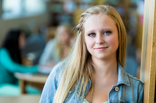 Beautiful high school senior smiles as she stands in doorway in the school library. She is leaning against the door. She has long blond hair and is wearing a denim jacket. Students are studying in the background.