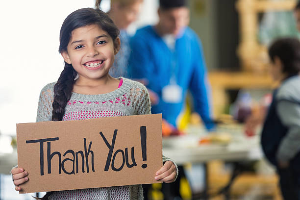 Pretty Hispanic girl holds 'Thank You!' sign in soup kitchen Cute Hispanic girl is holding a cardboard sign with 'Thank You!' witten on the board. She and her family are in a soup kitchen or food bank. She is smiling at the camera. Her brown hair is in a braid. Volunteers ar serving her family in the background. Focus is on the girl and the sign. thank you phrase stock pictures, royalty-free photos & images