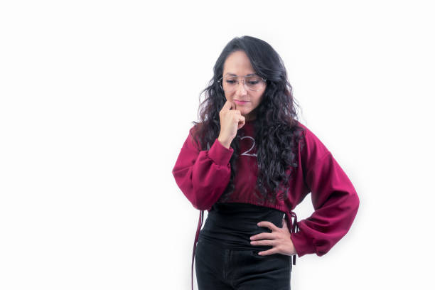 Pretty Hispanic Latina woman with thick long hair hunched over on white background indoors making expressions and gestures, pensive, wondering looking down with hand on face and mouth with hand on waist stock photo