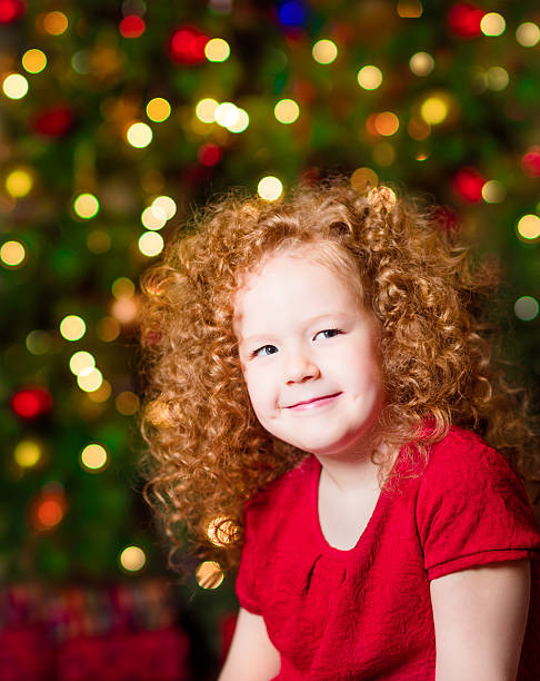 Pretty red-haired little girl wearing red dress sitting in front of...