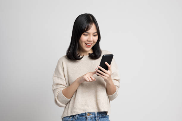 Pretty young asian woman using smartphone standing on isolated white background. She texting on cell phone. stock photo