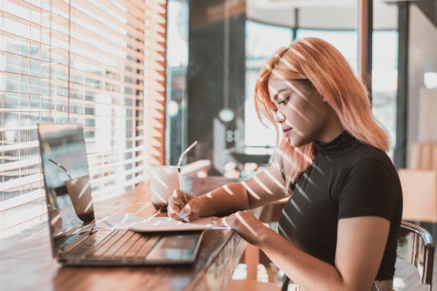 A pretty young focused professional asian woman writing down notes or making a draft. Office worker, businesswoman or freelancer at a coffee shop. stock photo