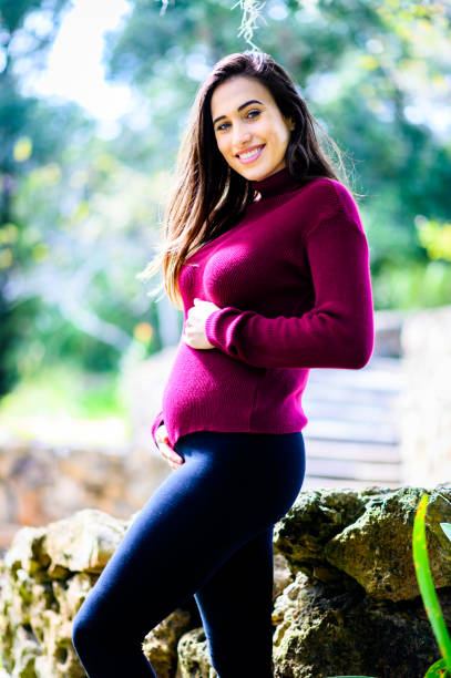 Pretty young pregnant Latinx woman cradles her 3 months pregnant belly in a casual happy portrait in scenic outdoor nature area stock photo