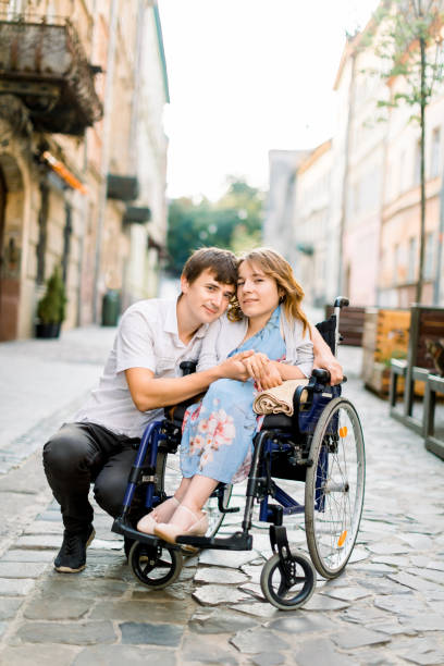 pretty young smiling woman in the wheelchair and handsome man looking at camera, holding hands, in love, walking in the old city center. - wheelchair street imagens e fotografias de stock