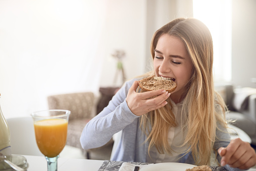 Pretty young teenage girl taking a bite of a healthy brown wholegrain roll as she enjoys a healthy breakfast at home with a glass of fresh orange juice
