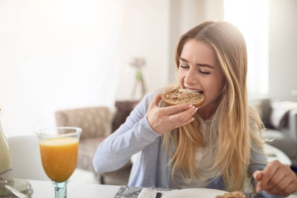 Pretty young teenage girl taking a bite of a roll Pretty young teenage girl taking a bite of a healthy brown wholegrain roll as she enjoys a healthy breakfast at home with a glass of fresh orange juice bun bread stock pictures, royalty-free photos & images