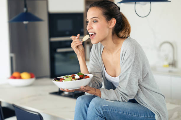 bella giovane donna che mangia insalata mentre è seduta in cucina a casa. - bella ragazza vegetariana foto e immagini stock