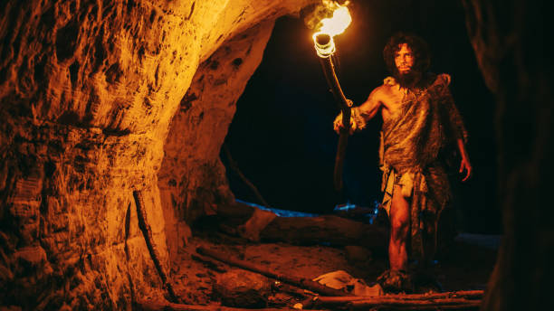 primeval caveman wearing animal skin exploring cave at night, holding torch with fire looking at drawings on the walls at night. neanderthal searching safe place to spend the night - fire caveman imagens e fotografias de stock