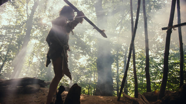 urzeitliche höhlenmensch tragen tierhaut hält stein gekippt speer, steht am höhleneingang blick über prähistorischen wald bereit, tier beute zu jagen. neandertaler auf der jagd im dschungel - neandertaler stock-fotos und bilder