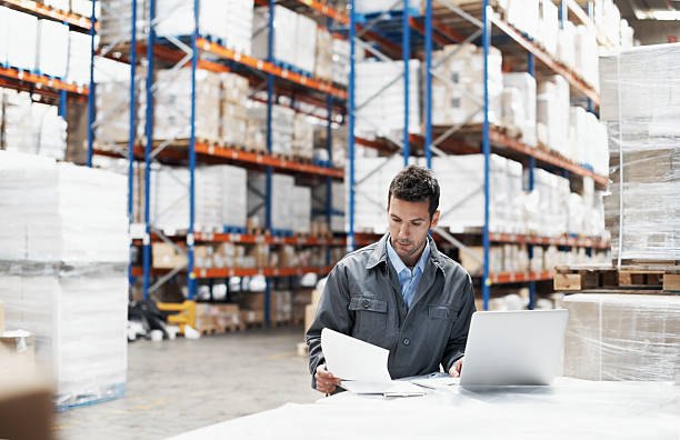 Prioritising the deliveries A young man using his laptop and looking over his notes while working in a warehouse factory photos stock pictures, royalty-free photos & images
