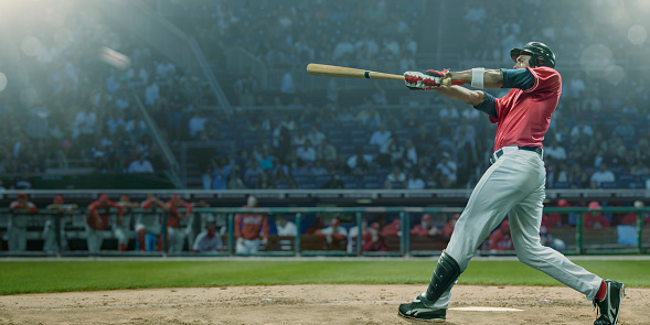 A professional male baseball player in mid swing with baseball bat outstretched having hit a baseball during a game. The player is dressed in generic red shirt and white trousers, and is wearing safety hemet and leg protector. He is standing in front of the dugout and crowd of spectators.