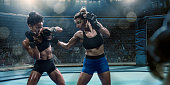 Two professional female mixed martial arts fighters competing in an octagon inside an indoor floodlit arena. Both fighters are in side on stance, and dressed in tight shorts and sports bras. One fighter has attacked with a jab or uppercut which has been blocked by her opponent.