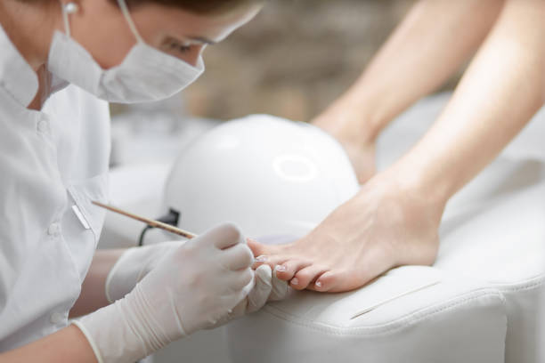 Professional in beauty salon cleaning nails on feet Close up of professional cleaning cuticle around nails on feet, with wooden stick. Female specialist in mask on face, making pedicure for client with uv lamp near. Beauty salon and service. pedicure stock pictures, royalty-free photos & images