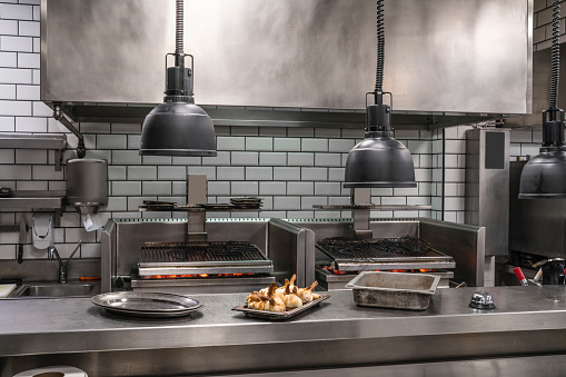Professional restaurant stainless steel kitchen with coal embers grill and roasted garlic tray in foreground