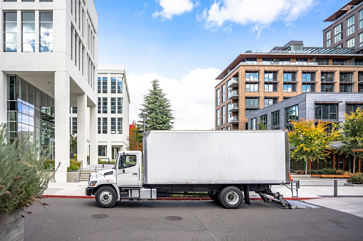 White middle class relocation rig day cab semi truck tractor with long spacious box trailer standing on the urban city street with multilevel apartment and office buildings unloading delivered goods
