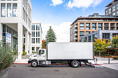 istock Profile of day cab medium size semi truck with long box trailer unloaded delivered goods to new multi-level apartments in unban city area 1351229888