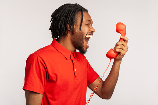 Profile portrait of male wearing red casual style T-shirt, screaming and yelling talking retro landline phone, complaining on connection quality. Indoor studio shot isolated on gray background.