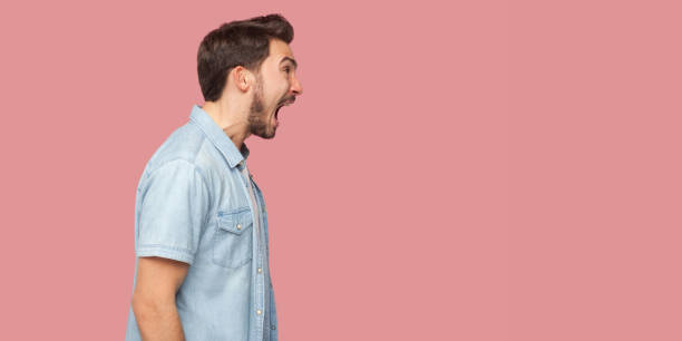 Profile side view portrait of angry or shocked handsome bearded young man in blue casual style shirt standing, looking forward and screaming. Profile side view portrait of angry or shocked handsome bearded young man in blue casual style shirt standing, looking forward and screaming. indoor studio shot, isolated on pink background. mouth open stock pictures, royalty-free photos & images