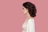 Profile side view portrait of calm beautiful brunette young woman with curly hairstyle in casual style standing with serious face and looking. indoor studio shot isolated on pink background.
