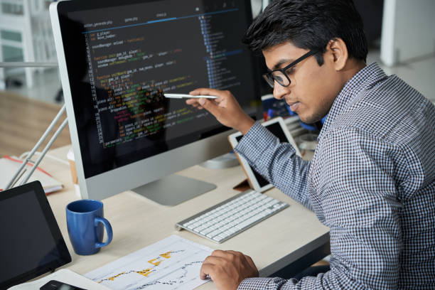 Programmer controlling the statistics of the site Serious Indian programmer in eyeglasses pointing at computer monitor and checking the statistics of website in document while working at office developer stock pictures, royalty-free photos & images