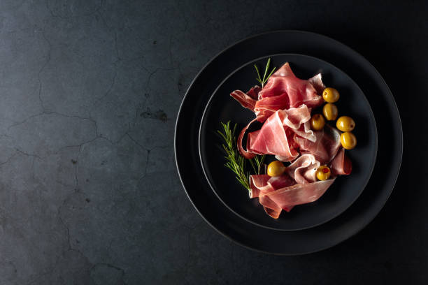 Prosciutto with rosemary and green olives on a black plate. Top view. Prosciutto with rosemary and green olives on a black plate. Top view, copy space. prosciutto stock pictures, royalty-free photos & images