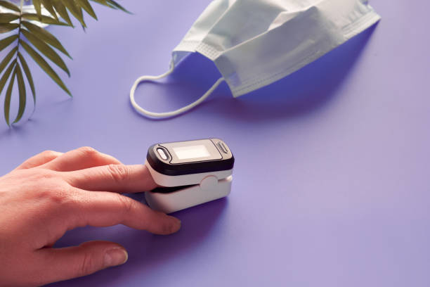 pulse oximeter. device monitoring oxygen saturation. reduced oxygenation is an emergency sign of covid-19 viral pneumonia. digital device on female finger, face mask, palm plant on purple neon table. - cor saturada imagens e fotografias de stock
