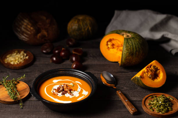 Pumpkin and chestnut soup with cream, pork belly and thyme aside an open pumpkin, chestnuts and bread on a vintage dark wood background. stock photo