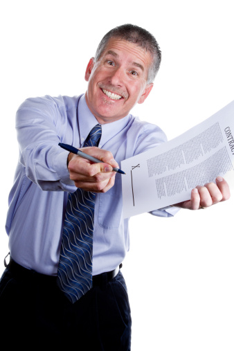 Smiling man with a cheesy grin gesturing for you to sign a contract, isolated on white. Man is mid 40s caucasian, has a mustache and short grey hair and is wearing a blue long sleeve shirt and dark blue tie. He is portraying a role as a used car salesman or some other type of high pressure sales.