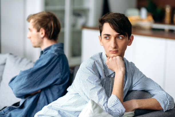 Quarrel between a gay couple in love. An upset guy and his partner are offended and ignore each other while sitting on the couch, turning away in different directions stock photo