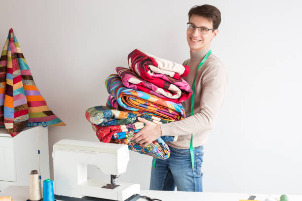 quilting and patchwork at workshop of tailor male - smiling tailor in glasses with a stack of stitched fabrics is near the desktop with a sewing machine, the neck hangs tailor measuring tape stock photo