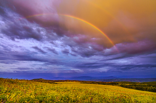 Rainbow Mountain Sunset Landscape Scenic View - Open meadow with colorful vibrant rainbow in sky with distant mountain views under dramatic sunset light.