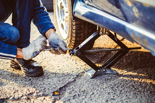 Jacking up a car to change a tyre after a roadside puncture with the hydraulic jack inserted under the bodywork raising the vehicle and the spare wheel balanced on the side.