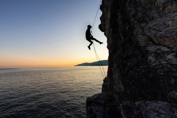 Rappelling down a Cliff during Sunset Silhouette of a Unrecognizable man rappelling down a steep cliff on the rocly ocean coast during a sunny summer sunset. Taken in Lighthouse Park, West Vancouver, British Columbia, Canada. west vancouver stock pictures, royalty-free photos & images