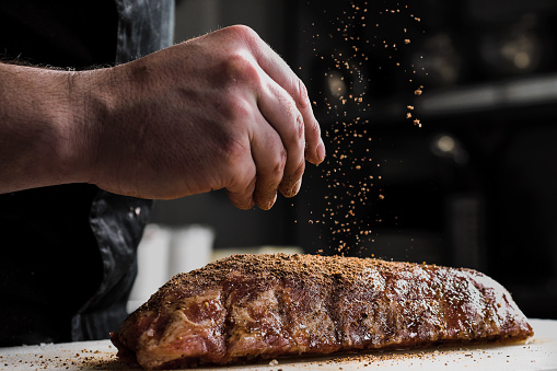 Raw piece of meat, beef ribs. The hand of a male chef puts salt and spices on a dark background, close-up.