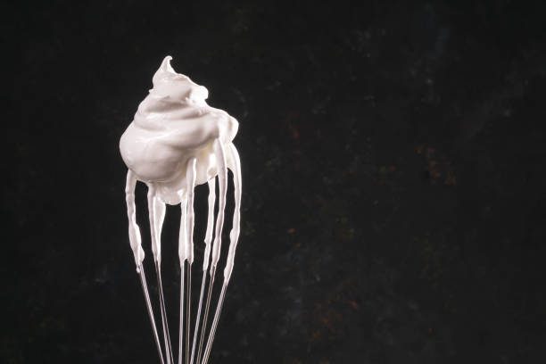 Raw uncooked meringue cream on whisk, dark background. Close up of a hand balloon whisk with beaten egg white raw meringue for perfect peaks on black background. Baking concept. Whipped cream, cookie recipe, copy space, studio shot. pavlova dessert photos stock pictures, royalty-free photos & images