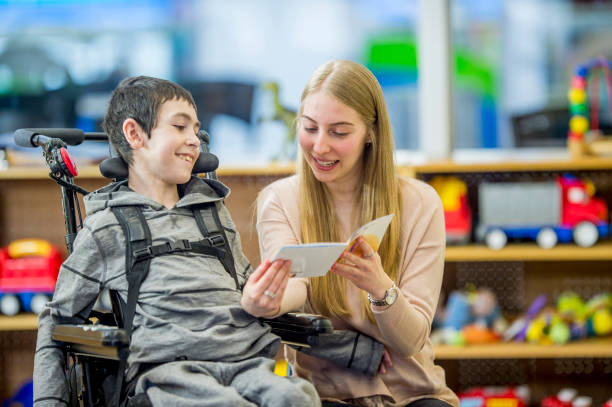 Reading A Story A boy in a wheelchair is indoors in his classroom. He is being assisted by his classroom helper. She is reading a story to him. physical disability stock pictures, royalty-free photos & images