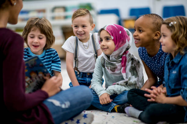 Reading A Storybook A multi-ethnic group of school children are indoors in a classroom. They are wearing casual clothing. They are sitting on the floor and eagerly listening to their teacher read a storybook. elementary age stock pictures, royalty-free photos & images