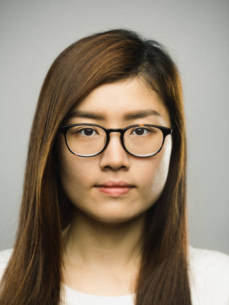 Real chinese young woman with blank expression Close up portrait of asian young woman with blank expression against gray background. Vertical shot of real chinese woman staring in studio with long brown hair and eyeglasses. Photography from a DSLR camera. Sharp focus on eyes. indonesian girl stock pictures, royalty-free photos & images