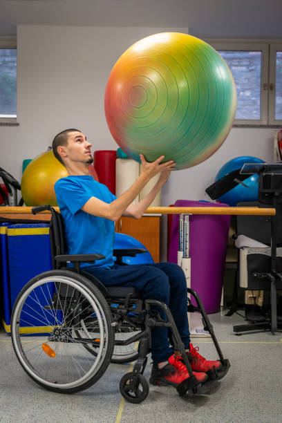 A real disabled skinny young man and wheelchair lifts weights and a ball on the rehabilitation of the disabled in the rehabilitation center. stock photo