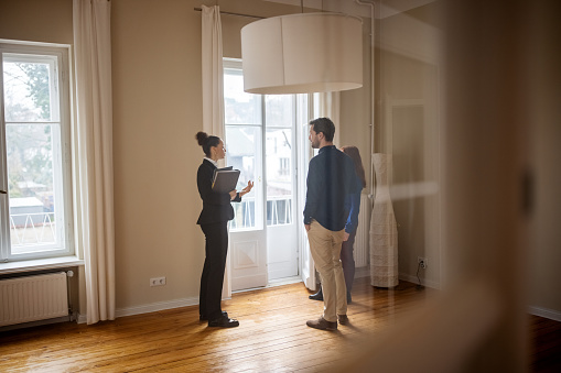 Full length of businesswoman discussing with customers at empty home. Female realtor is standing with man and woman by window. They are at new apartment.
