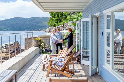 Real estate agent showing a mature couple a new house. The house is contemporary. All are happy and smiling and shaking hands. The couple are casually dressed and the agent is in a suit. Waterfront can be seen in the background
