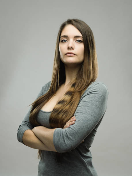 Real young woman studio portrait Portrait of confident beautiful young woman with neutral expression and arms crossed. Real people female is against gray background looking at camera. She has long brown hair. Vertical studio photography from a DSLR camera. Sharp focus on eyes. snob stock pictures, royalty-free photos & images