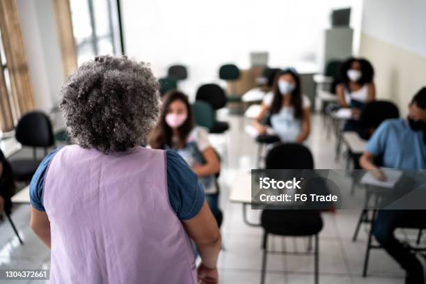 Rear view of a teacher in front of class teaching students - wearing face mask