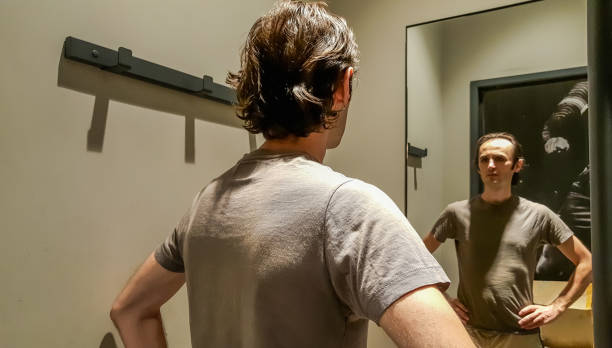 Rear view of a young Caucasian handsome man trying new clothes in front of a mirror in a changing room of a clothing store stock photo